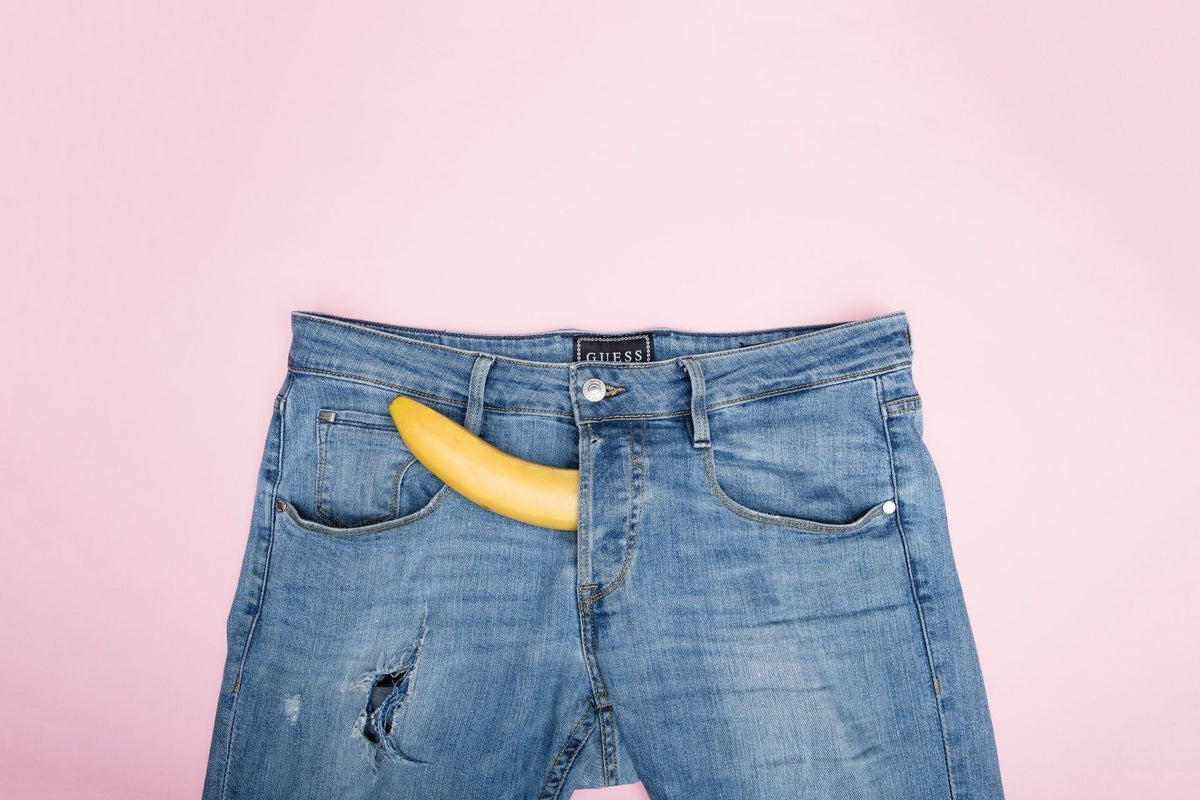 Banana sticking out right side of the opening on a pair of jeans upon a light pink background